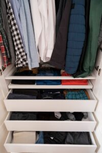 A photograph of a closet with clothes hanging above a set of drawers.